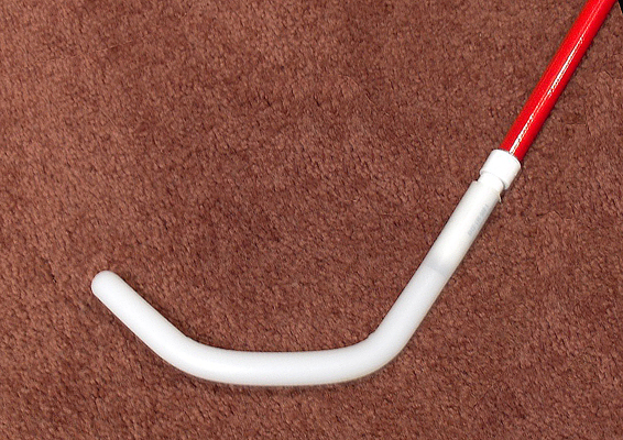 Photo shows the Bundu Basher Tip at the end of a cane.  The tip is long and cylindrical, the same diameter as the cane, and is about 10 inches long.  The first 4 inches extend straight from the end of the cane, then it bends about 45 degrees and continues for another 3 inches (this is the part that is horizontal to the ground).  It then bends again and the last 3 inches is turned up at a 45-degree angle from the ground (shaped like a wide 'U' or like a hockey stick with its tip bent upward).